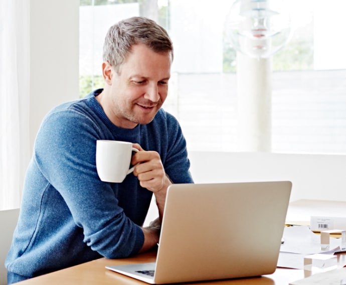 Bir adam evde dizüstü bilgisayar kullanarak kahve içiyor.
